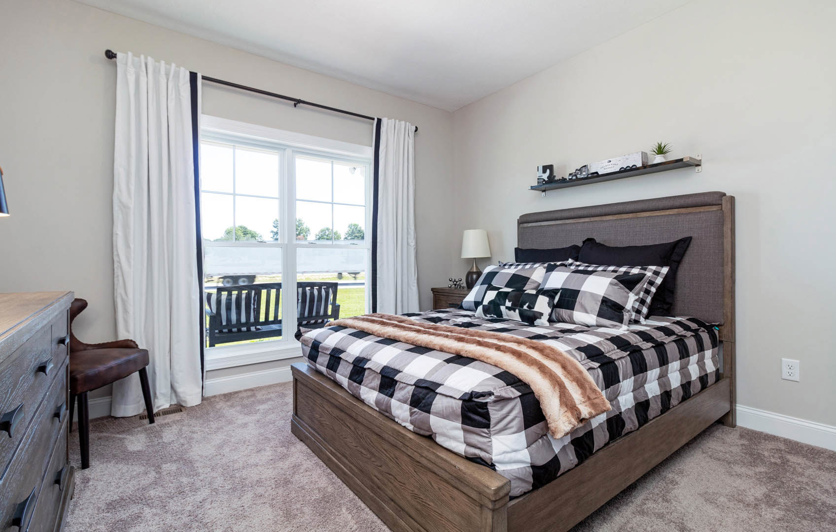 Plaid blanket and pillows on a bed beside a window with blue-striped white curtains, potted plant on a shelf, white outlet visible on wall, chair near bed.