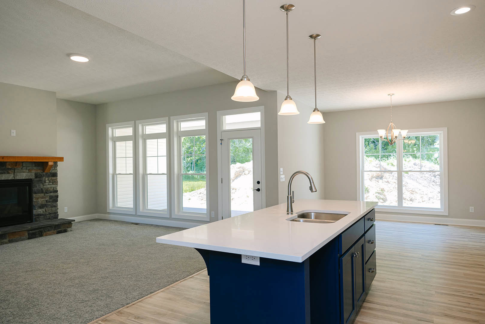 Blue kitchen island with white countertop, built-in sink and faucet, white cabinetry, stone fireplace wall, pendant light fixture near window, hardwood flooring