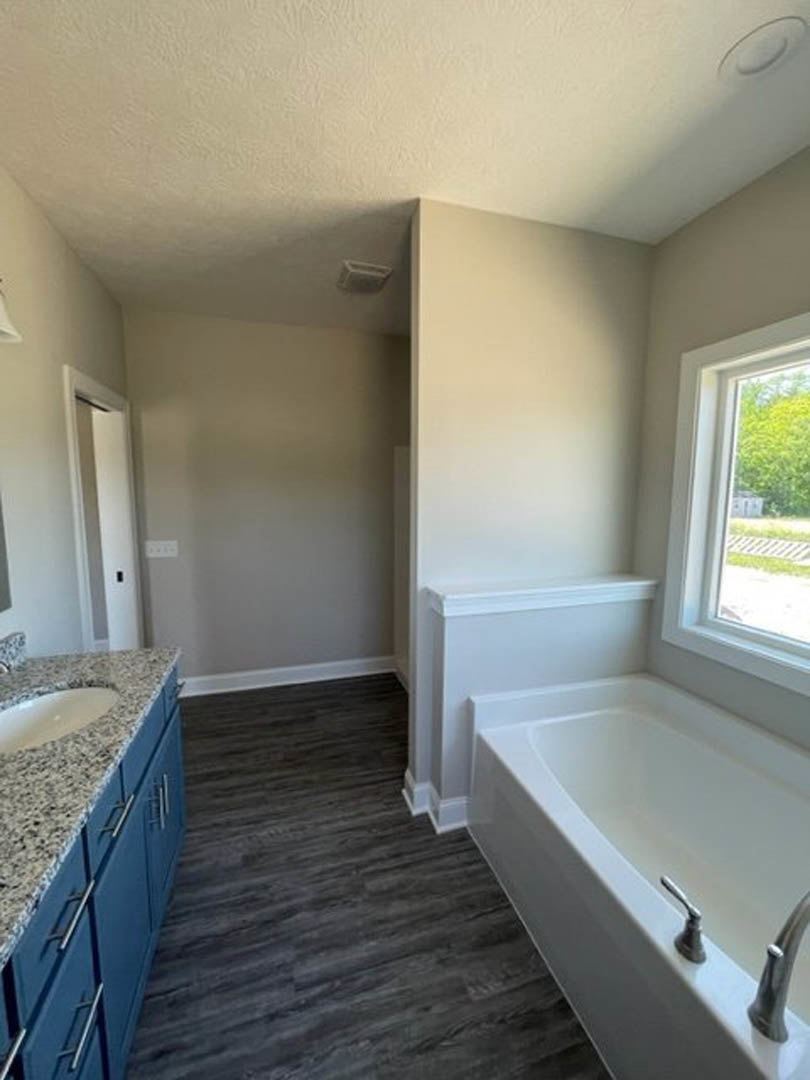 White freestanding bathtub with chrome faucet on dark wood floor, rectangular sink with modern tap, large window showing green tree outside