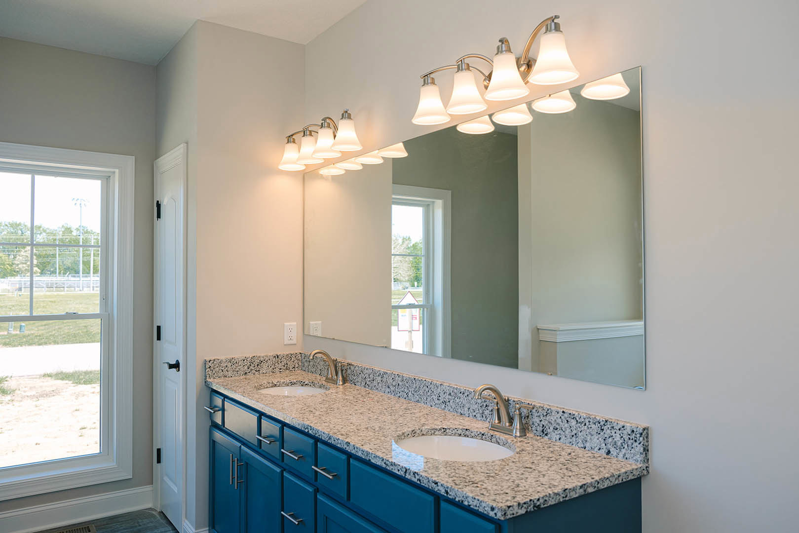 Bathroom with blue shaker cabinets, wide vanity mirror, white countertop, chrome faucet, window overlooking grassy yard and fence, row of modern light fixtures above mirror