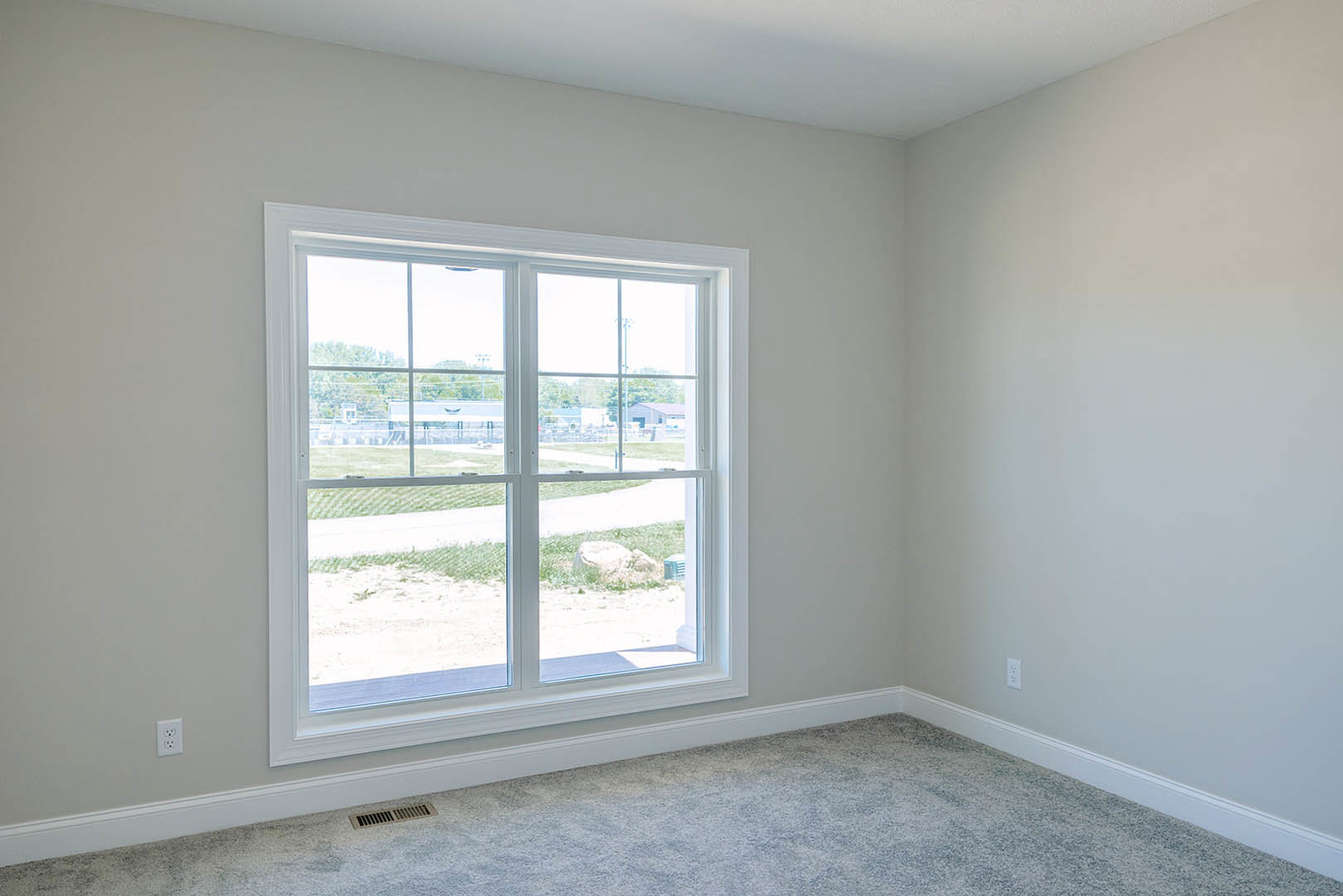 Neutral-toned carpeted floor in a room with white plaster walls, large window overlooking grassy field with a rock, window blinds partially drawn