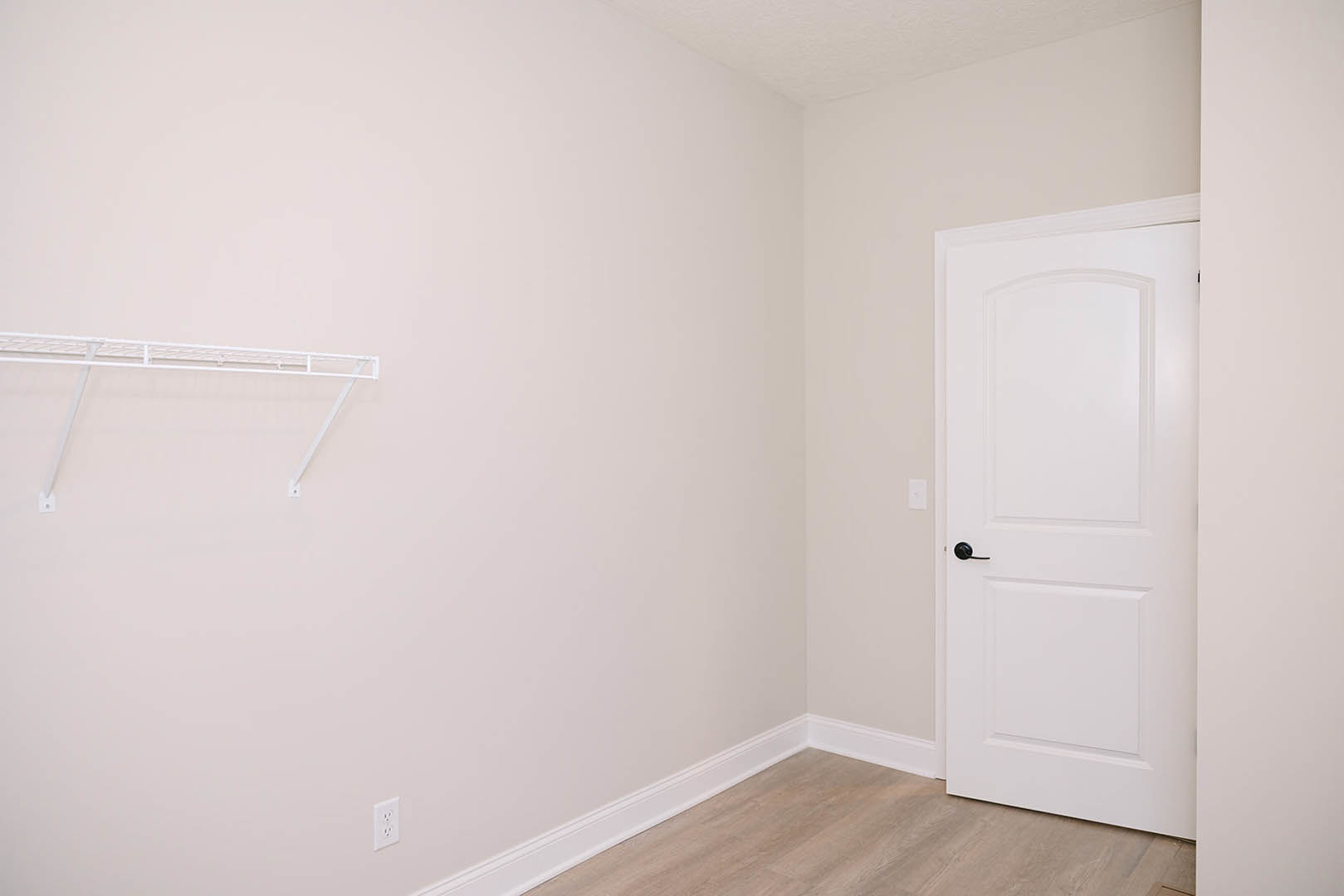 White door with black handle set in a room featuring wood flooring, white baseboards, white clothes rack, and white cabinet against white walls.