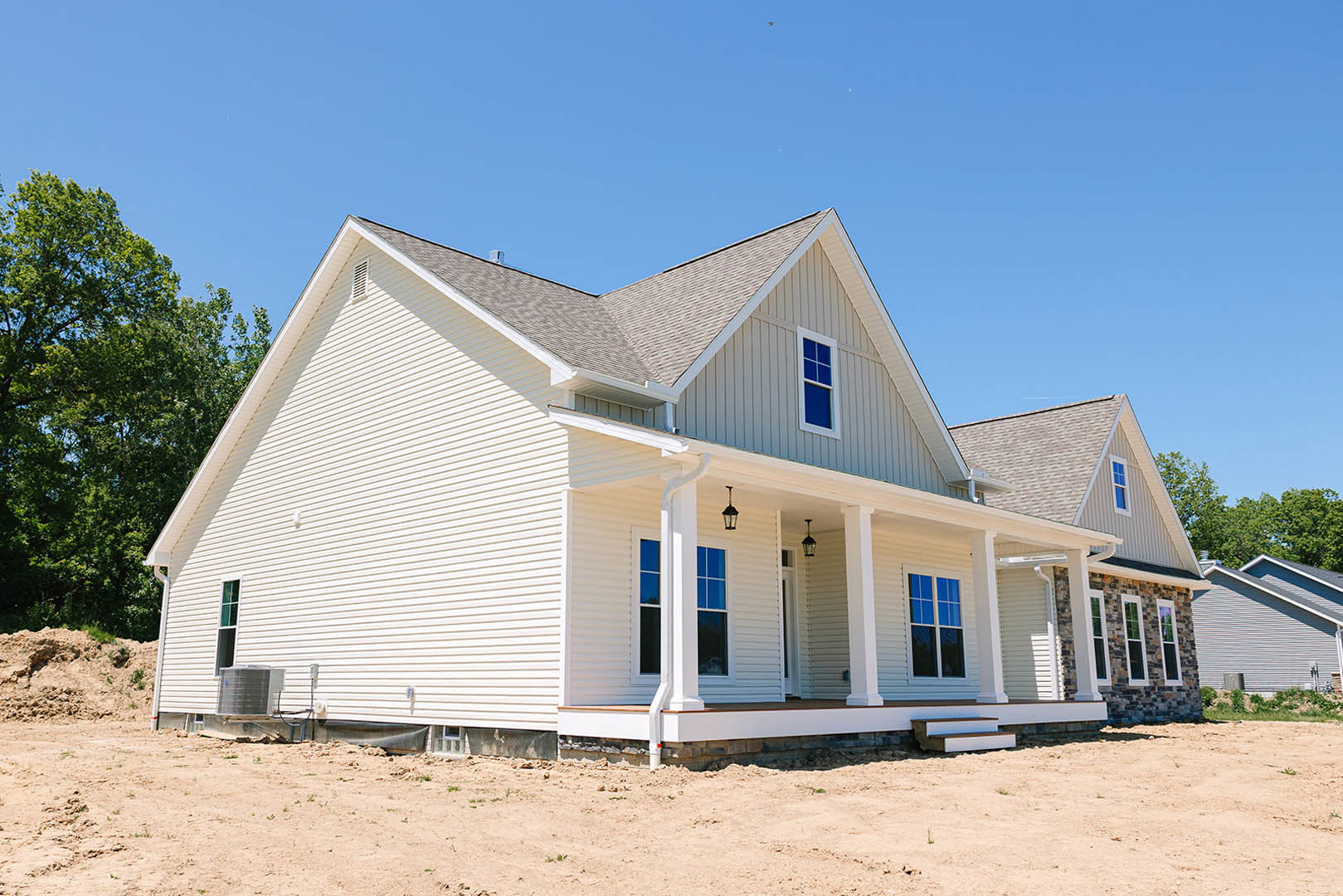 Two-story house under construction with exposed framing, white siding, covered porch, large windows, dirt yard with white pipe, tree nearby, air conditioning unit mounted, and