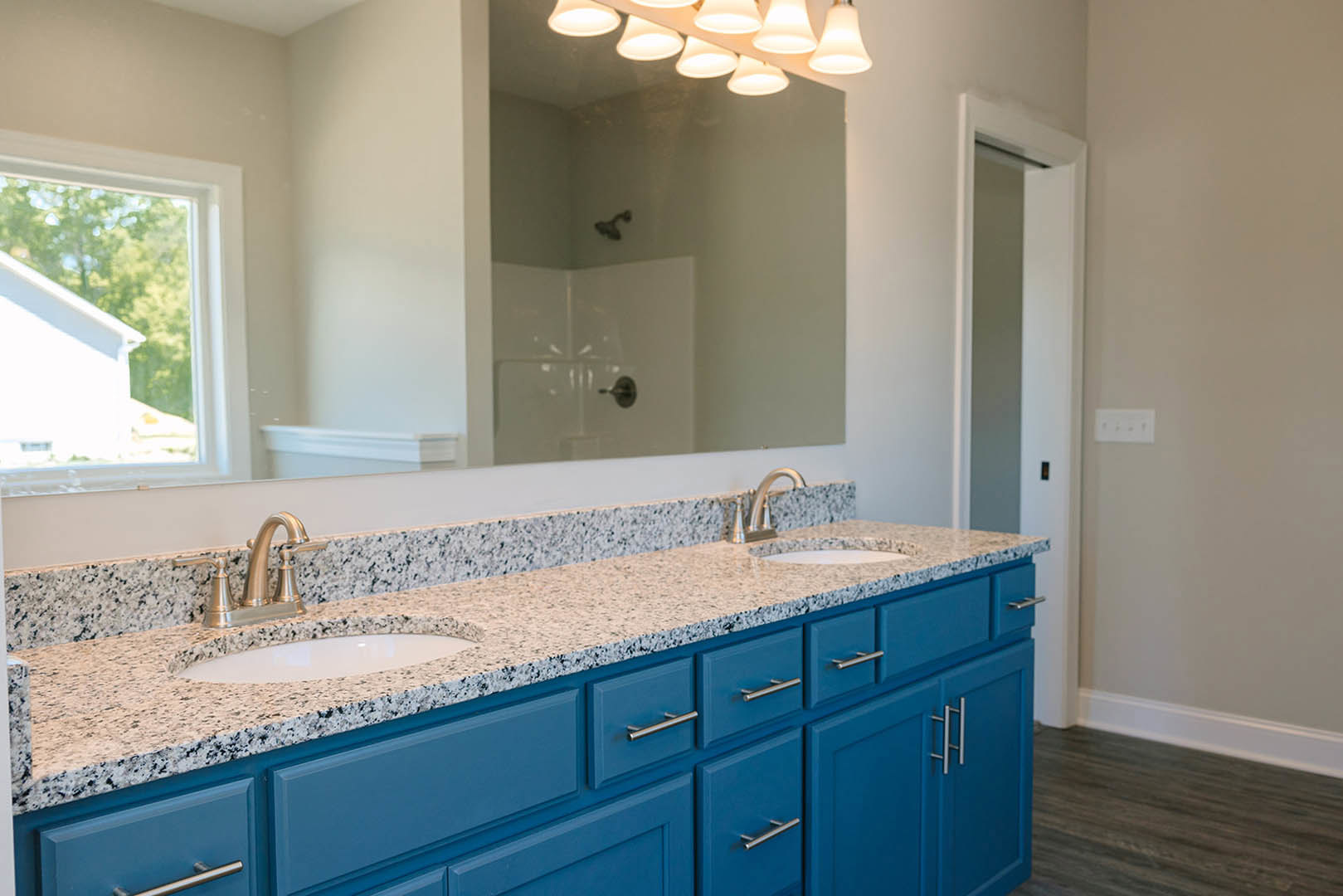 Blue bathroom vanity with white countertop and sink, rectangular mirror above, ceiling lights, white wall outlet with black switches, window showing trees outside, tile flooring
