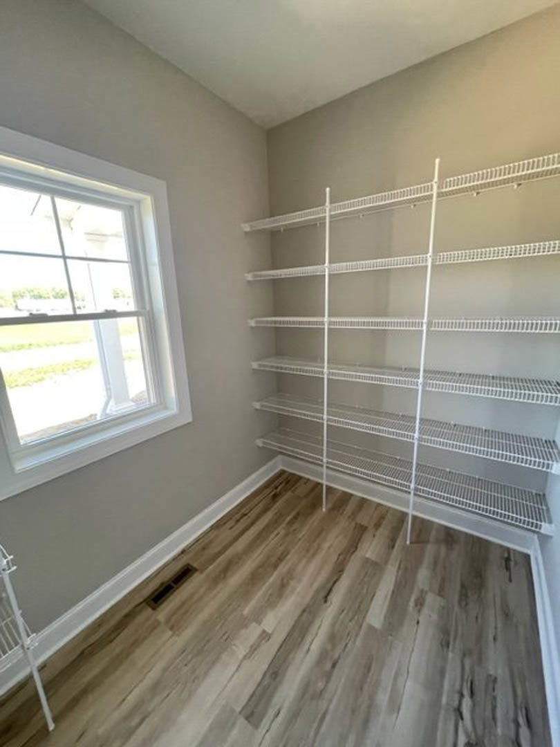 Empty closet with built-in white shelves, wood flooring, and a window with a white frame letting in natural light.