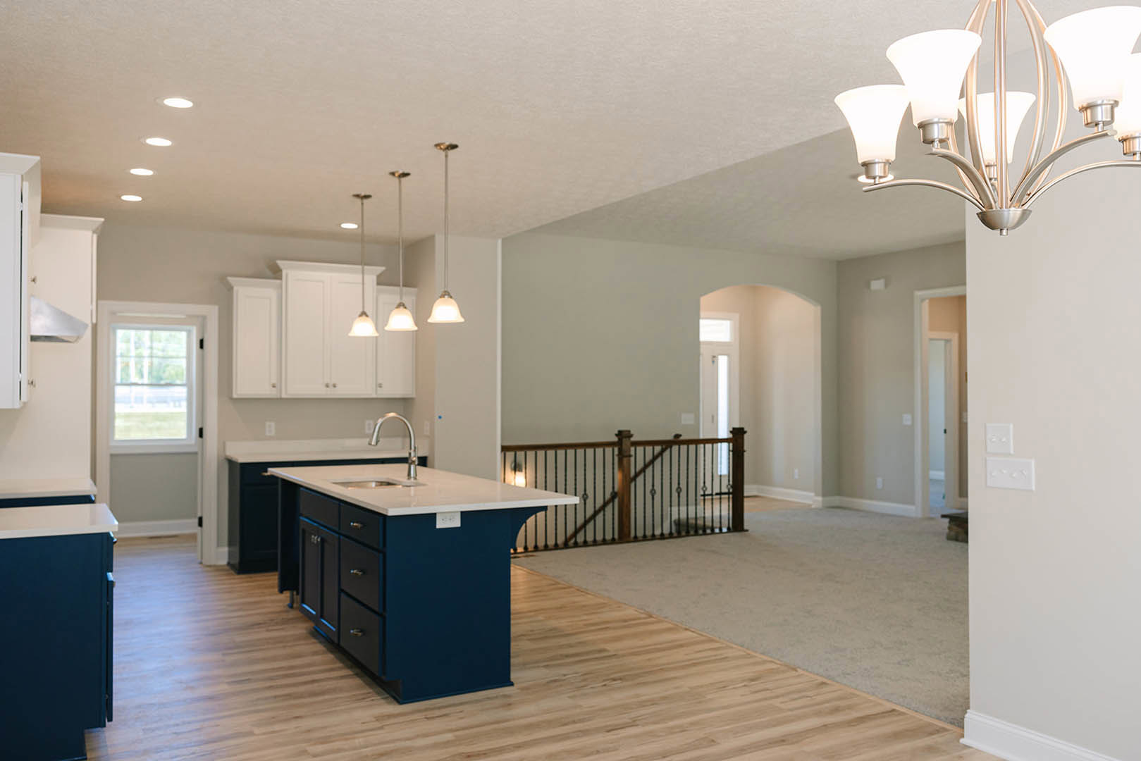 Open-concept kitchen and living room featuring wood flooring, white kitchen island with sink, wooden staircase railing, white dining table, modern chandelier, and cabinetry with