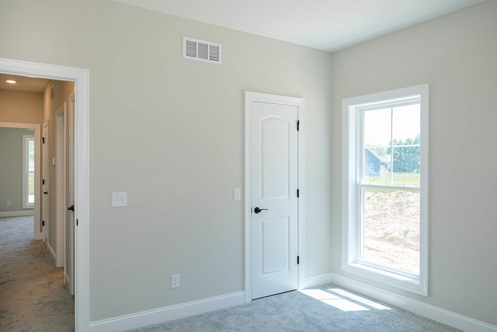 White carpeted floor, white walls with a white door featuring a black handle, window showing blue house exterior, white light switch and wall vent.