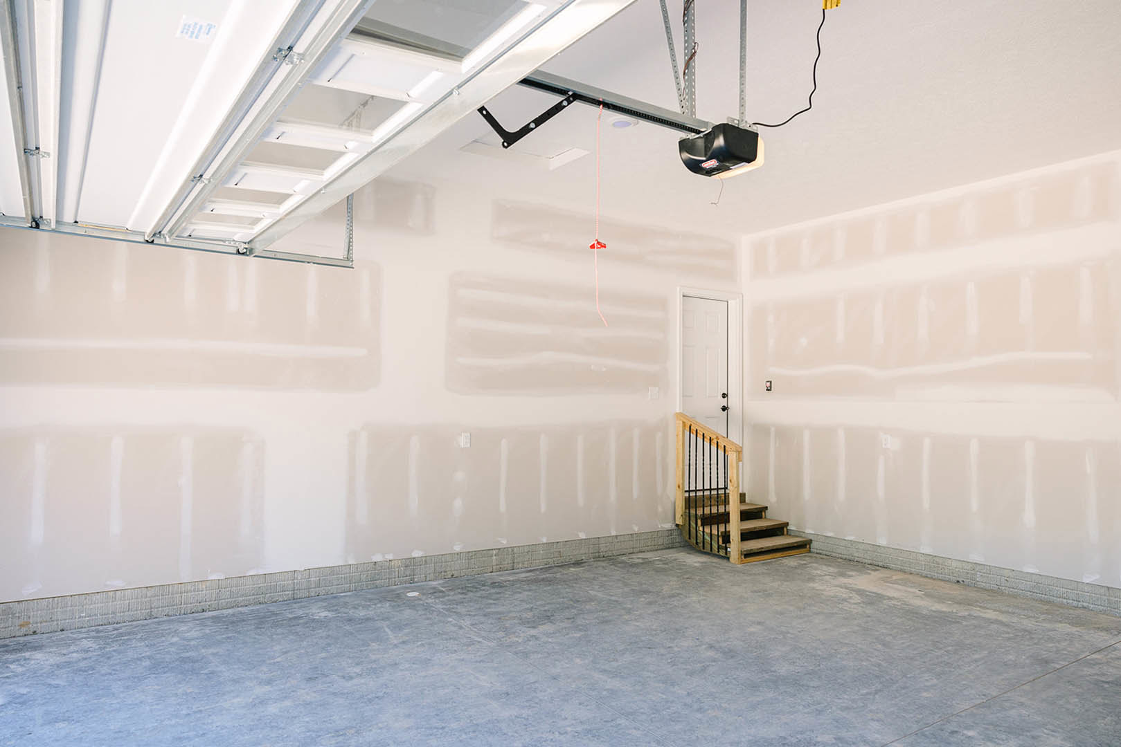 Wooden staircase with metal railing beside a white door, concrete floor, plaster ceiling with recessed fixture, black frame with visible screw.