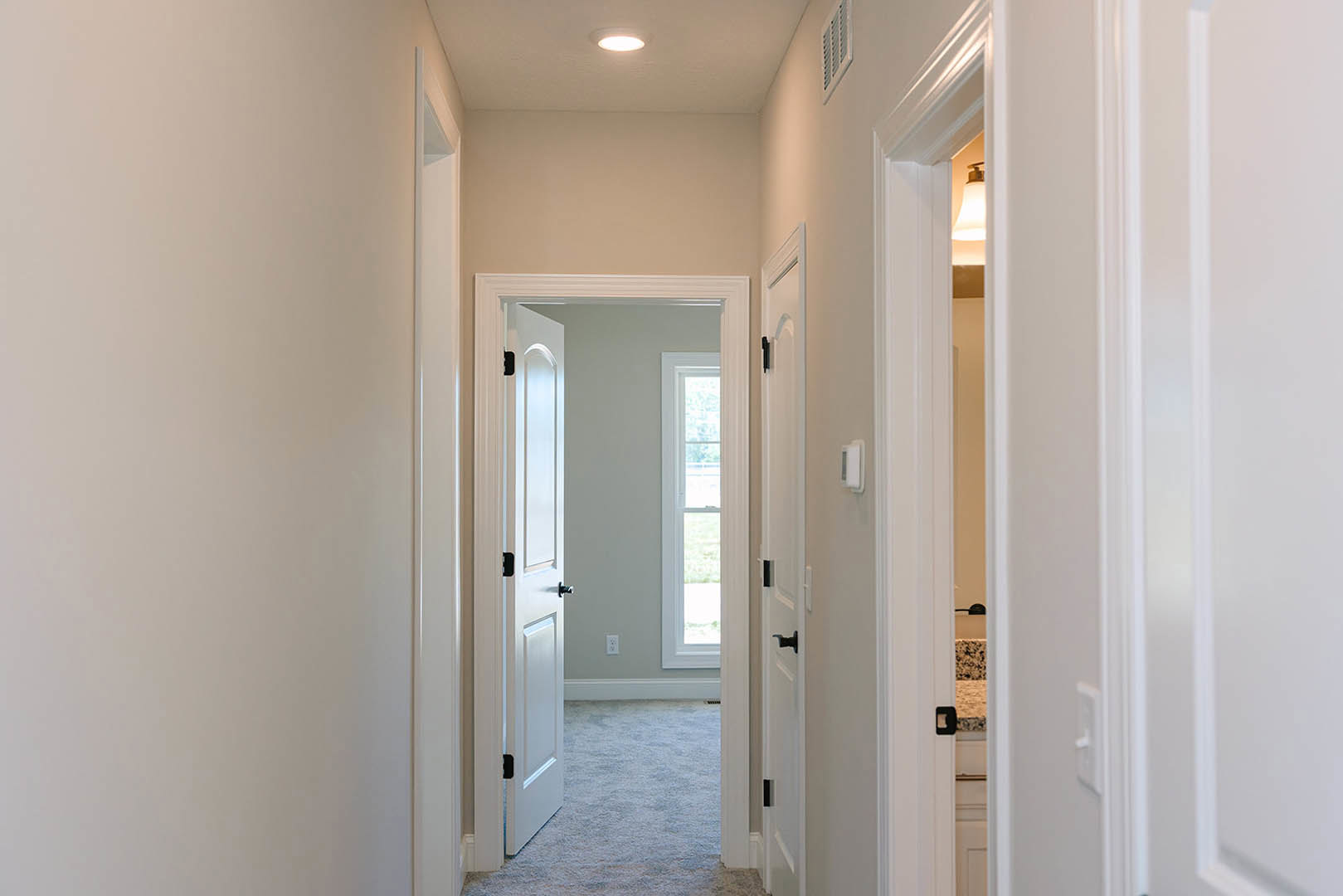 Hallway with white paneled doors, carpeted floor, white walls, and a window with a white frame; black rectangular object mounted on one door
