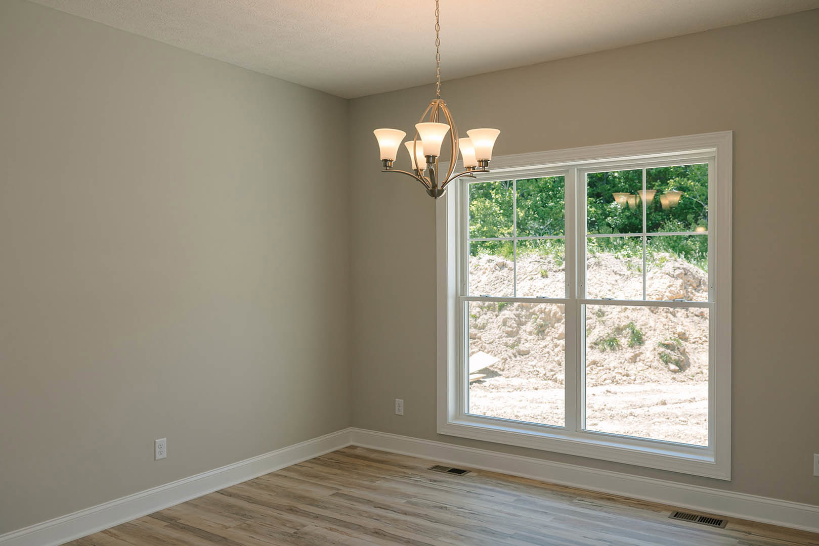 Chandelier hanging from ceiling above wood floor with vent, large window with white molding and blinds, plaster walls, partial view of table and lamp.