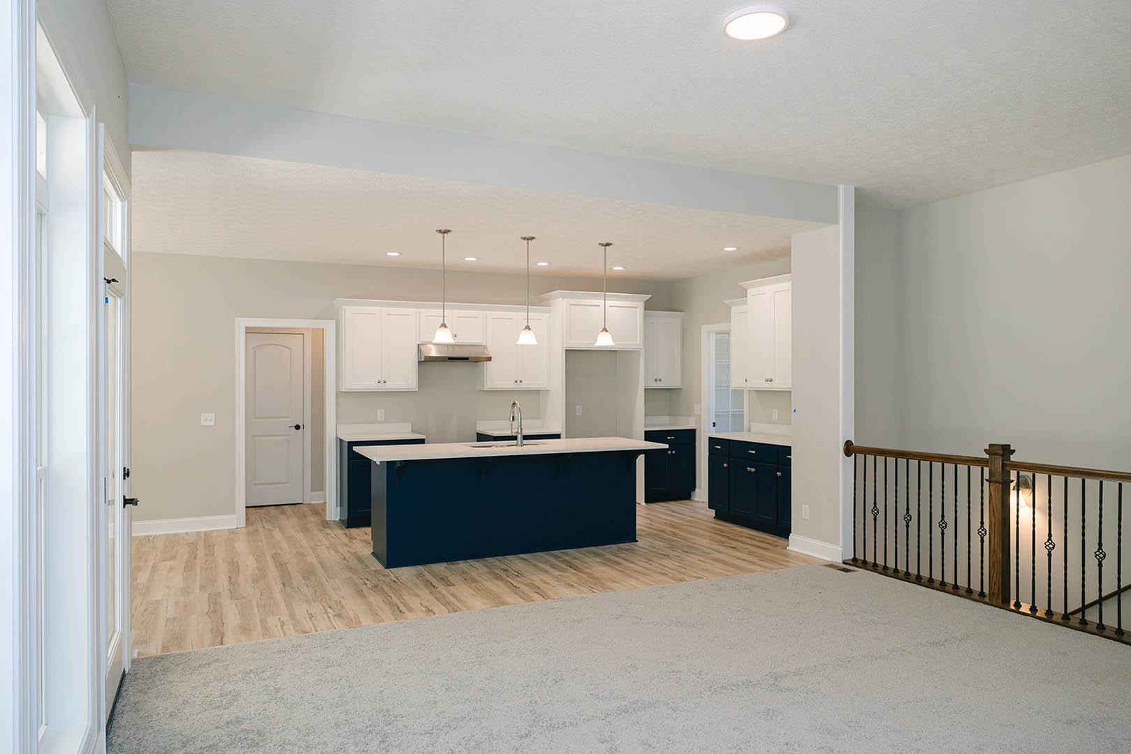 Open-concept kitchen and living room featuring blue countertop with sink, white cabinetry, wood flooring with carpet, modern ceiling light, black-handled white door, and metal
