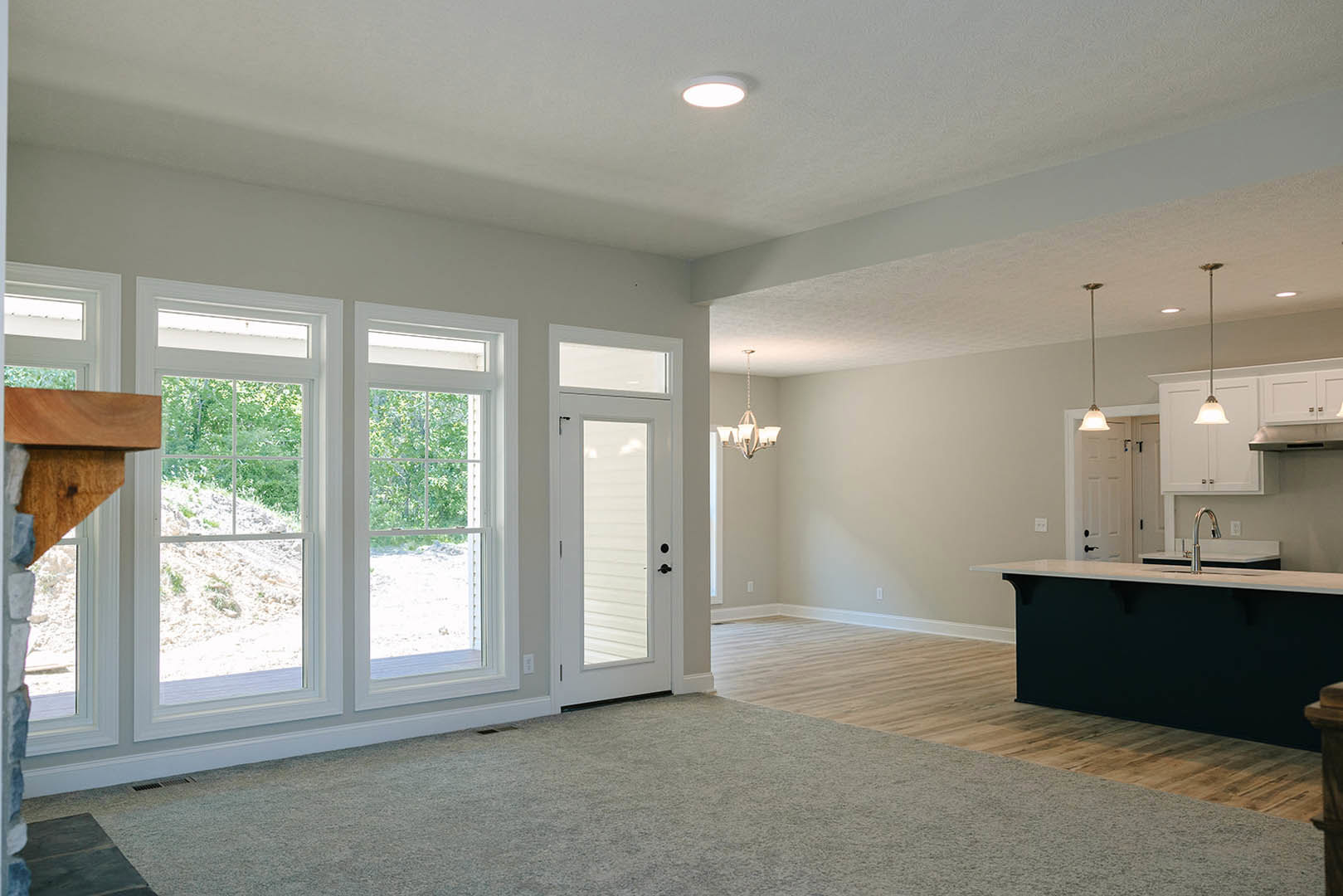 Open kitchen with black countertop and silver pendant lamp, adjacent dining table, white door with glass window, large window overlooking forest, light wood flooring, white