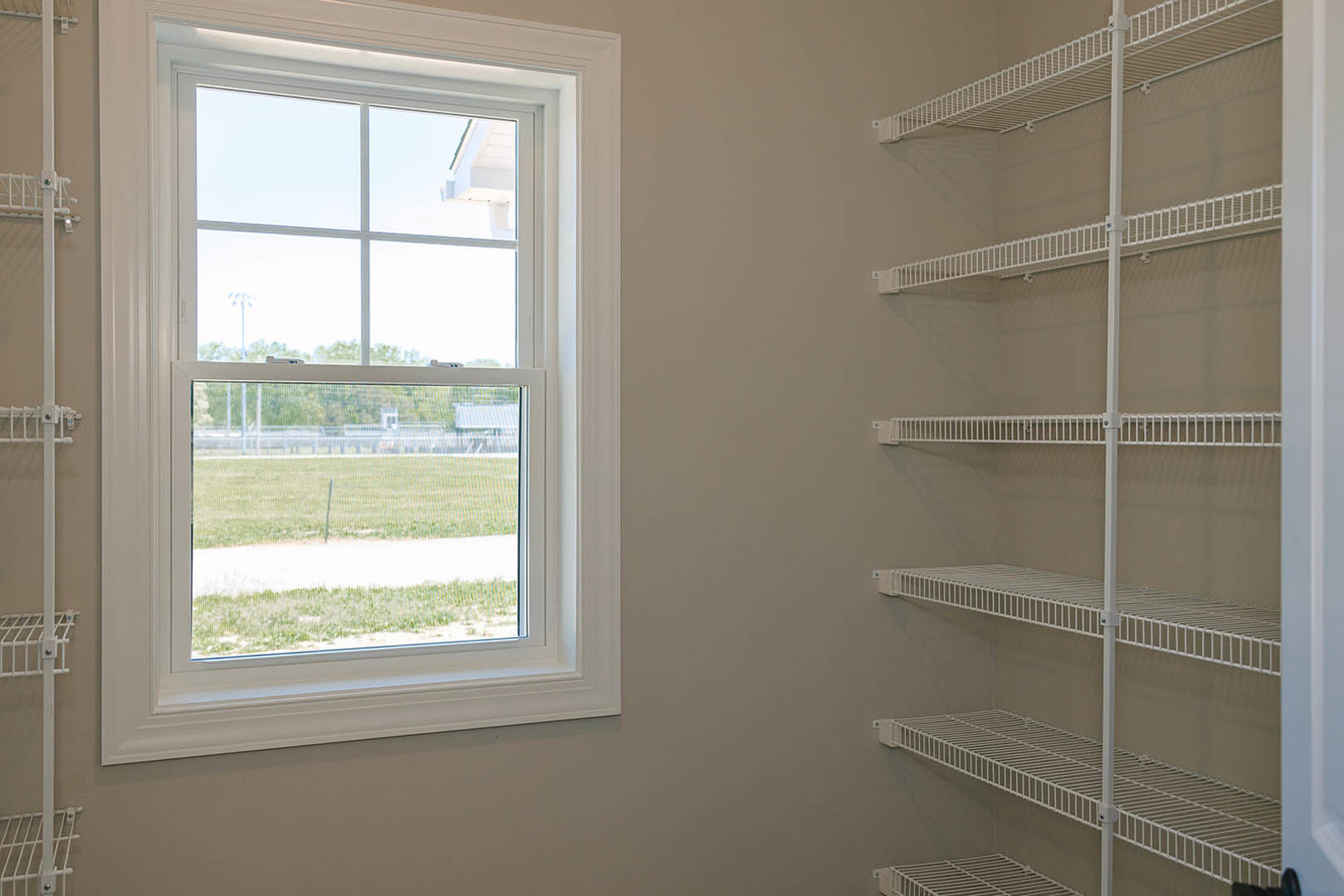 White built-in shelves with metal rods beside a large window overlooking a grassy field, set against light-colored walls in a modern interior.