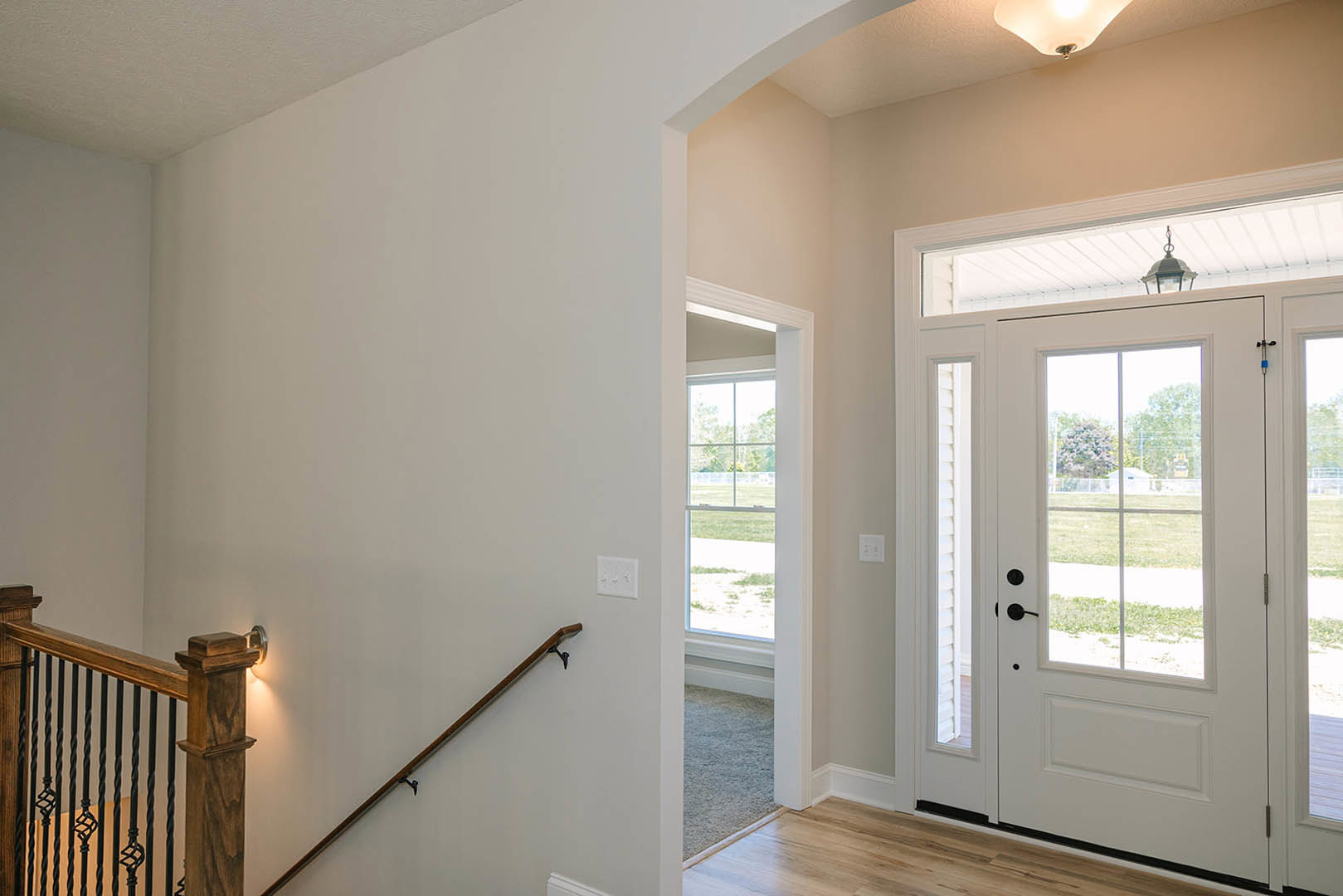 Hallway with grey carpet, white walls, staircase featuring a close-up of a wooden handrail, white door with glass panes, and a white light switch.