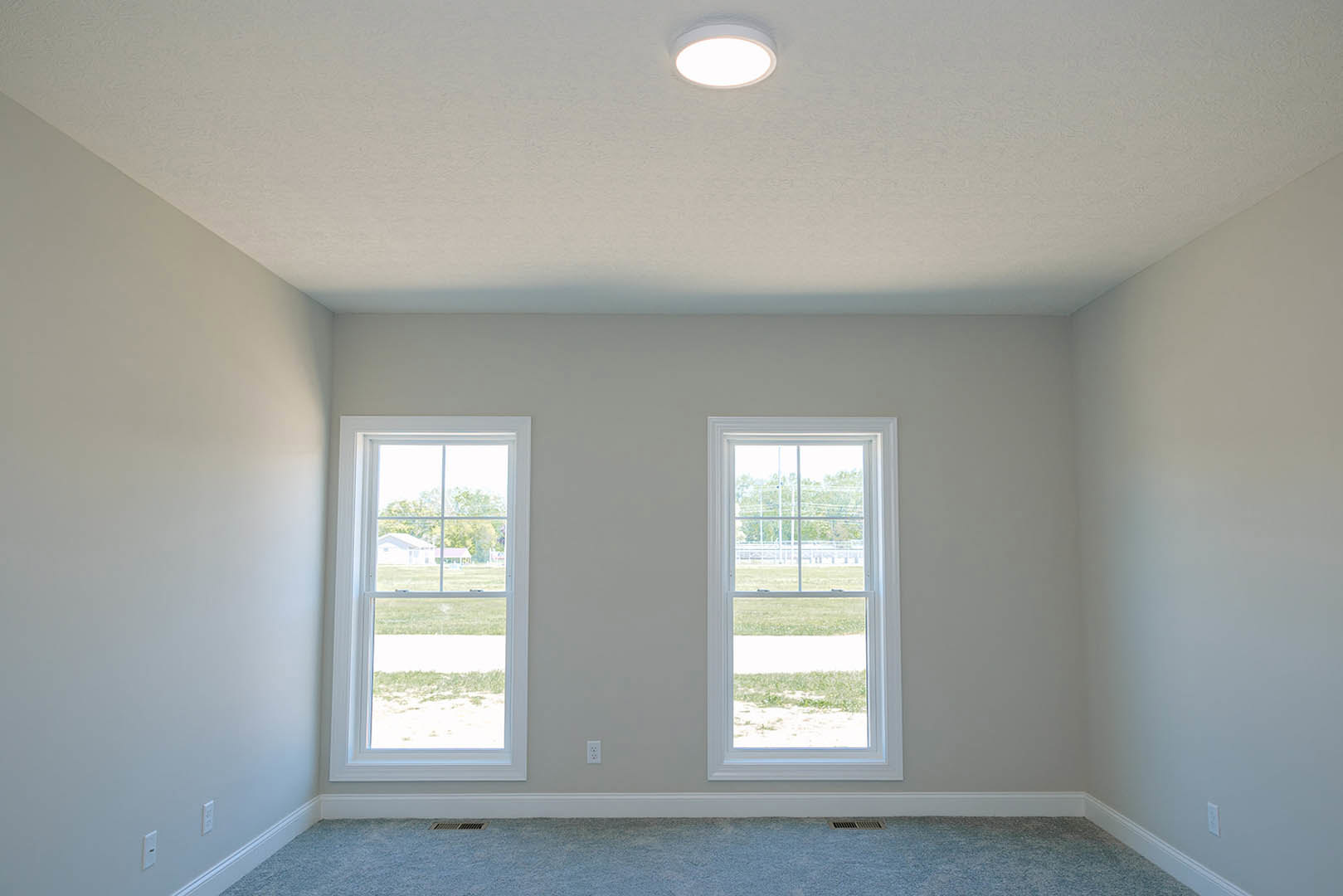 Carpeted room with two rectangular windows, white walls, ceiling light fixture, views of grassy yard and wooden fence