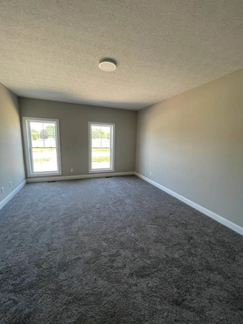 Bedroom with two rectangular windows, grey carpet flooring, white plaster walls, and ceiling; view of green grass outside.