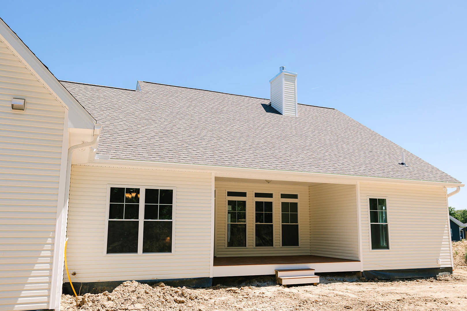 White siding exterior with multiple windows, white chimney, and bench in front of a custom home under construction beneath a clear blue sky