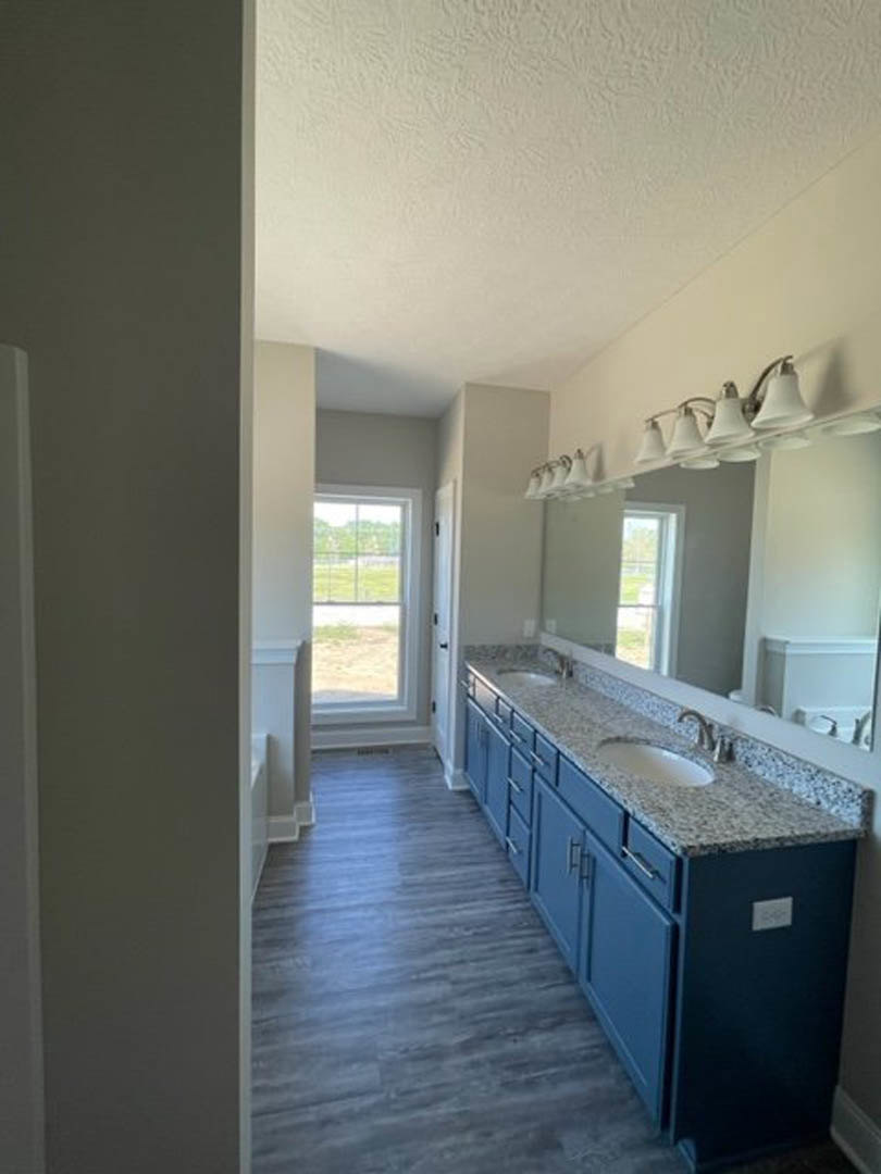 Bathroom with double sinks, marble countertop, large mirror, blue cabinetry, row of bell-shaped light fixtures, tile flooring, and window overlooking fenced yard