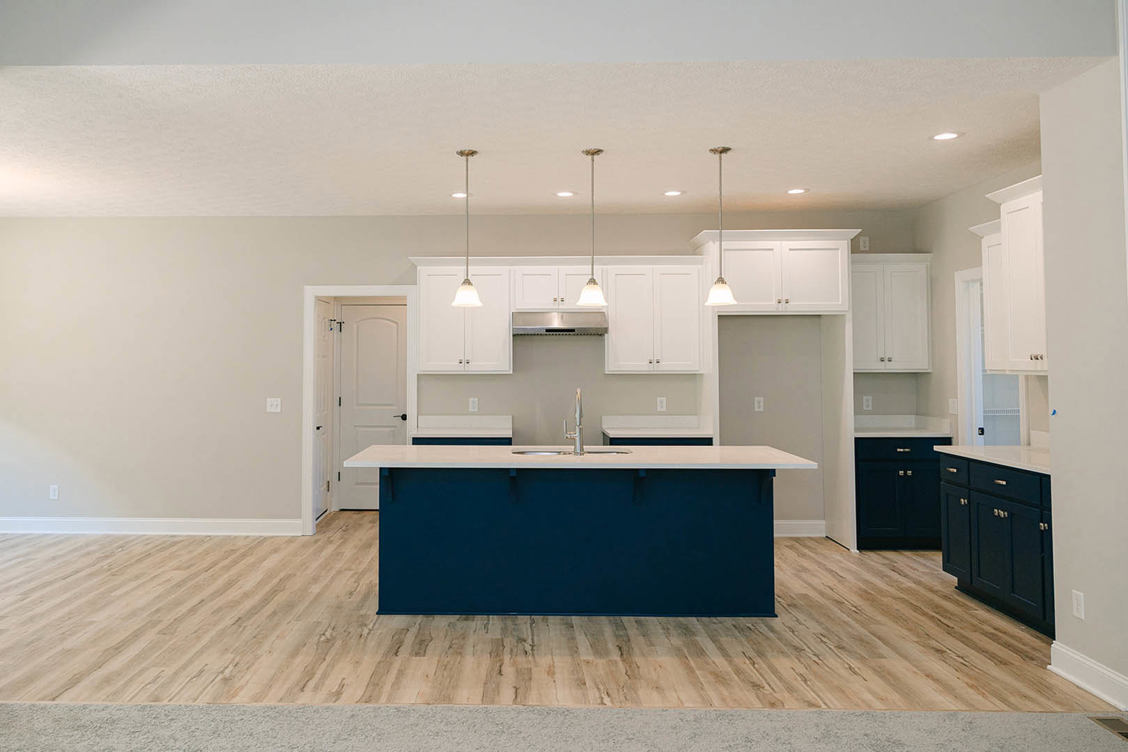 Kitchen with white shaker cabinets, blue island, wooden flooring, white door with black handle, stainless steel sink, and black cabinet drawer fronts.