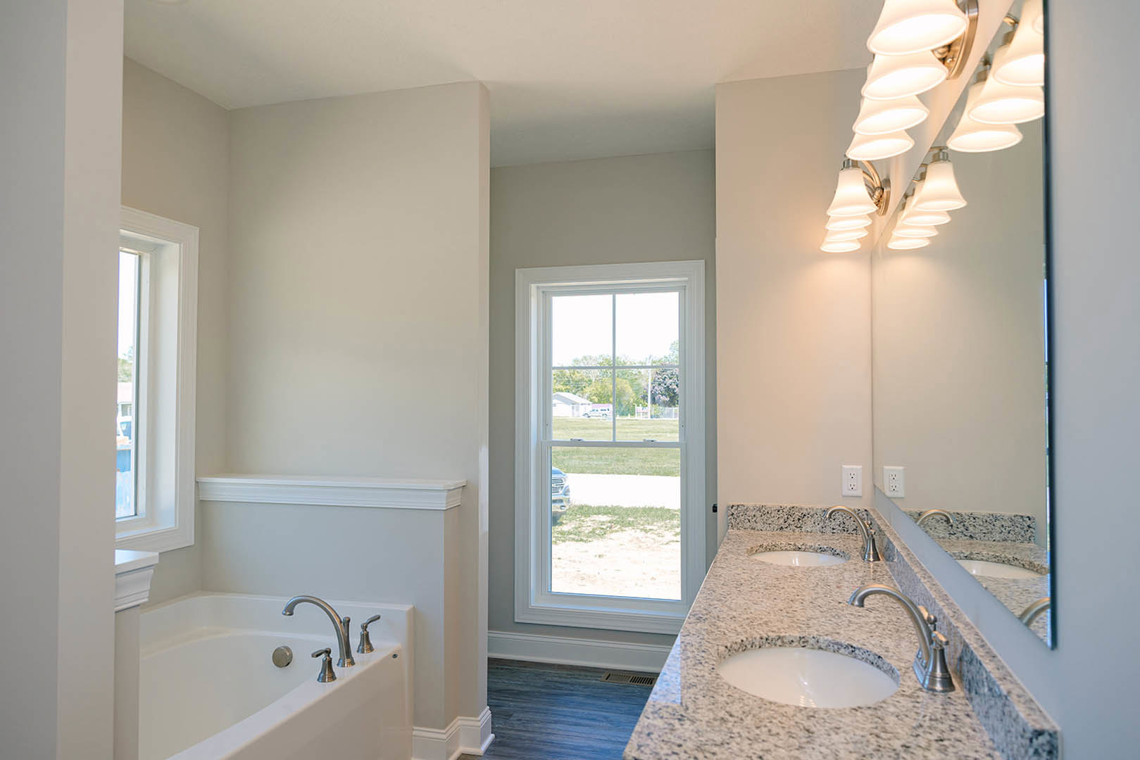 Bathroom with marble countertop sinks, freestanding bathtub, row of wall-mounted lights, large window showing parked car outside, blue wood floor with vent, mirrors above sinks