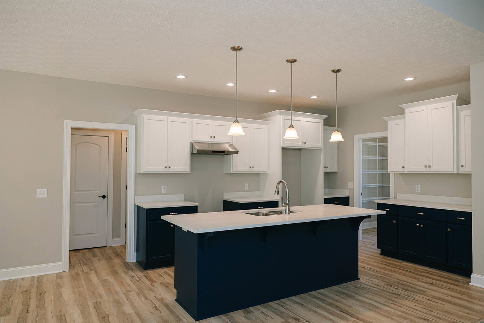 White kitchen cabinets, central island with quartz countertop, stainless steel sink, white door with black handle, recessed ceiling lights, hardwood flooring