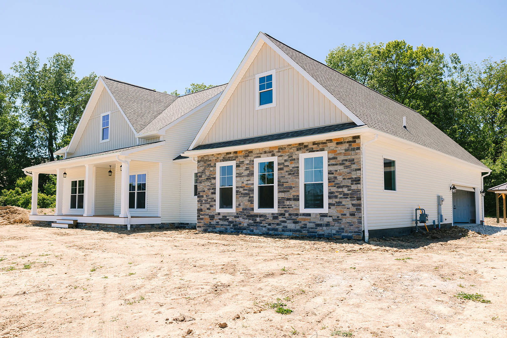White house with triangular roof, large front window, and unfinished dirt yard bordered by a brick wall; sparse landscaping and clear sky in background.