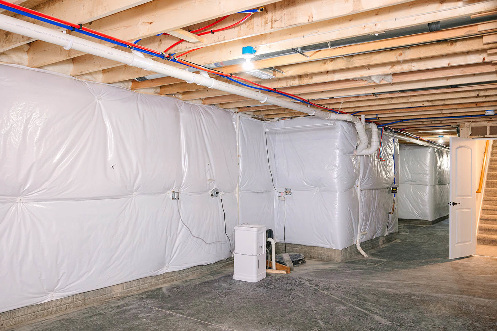 Basement room with white plastic wall coverings, exposed pipes and wires, white door with handle, staircase, and a rectangular object with black top; unfinished ceiling with beams.