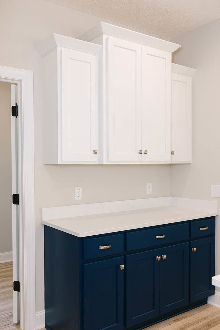Kitchen featuring white upper cabinets with silver knobs, blue lower cabinets and drawers, white countertops, stainless steel sink, and a black hinge on a white door.