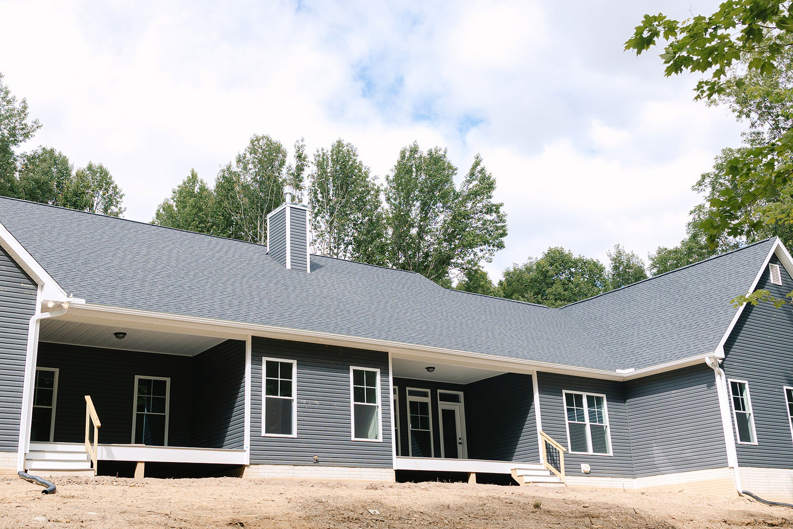 Two-story home with wide covered porch, white framed windows, gray siding, front steps, brick chimney, and mature trees in the background