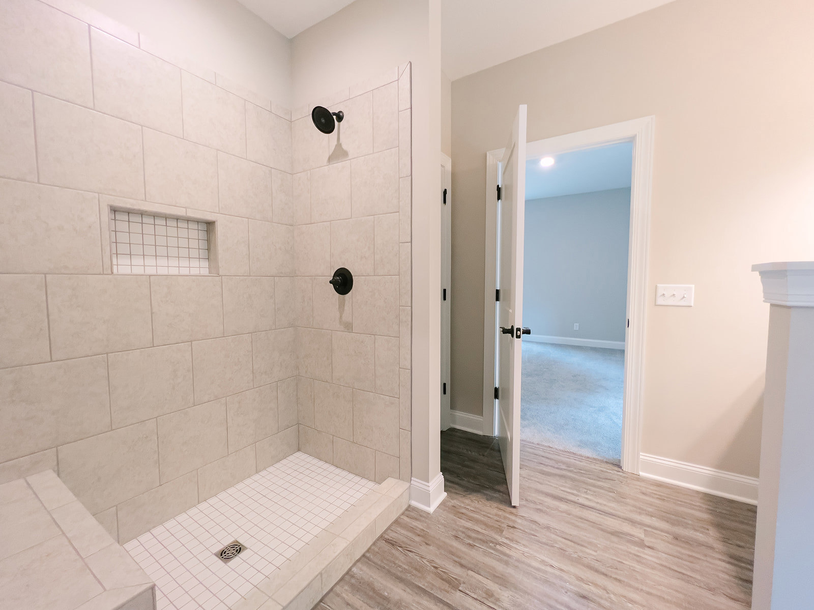 Bathroom with wood flooring, white tile shower walls with white border, open door leading to adjacent room, black round light fixture, white switch with knobs, shower floor