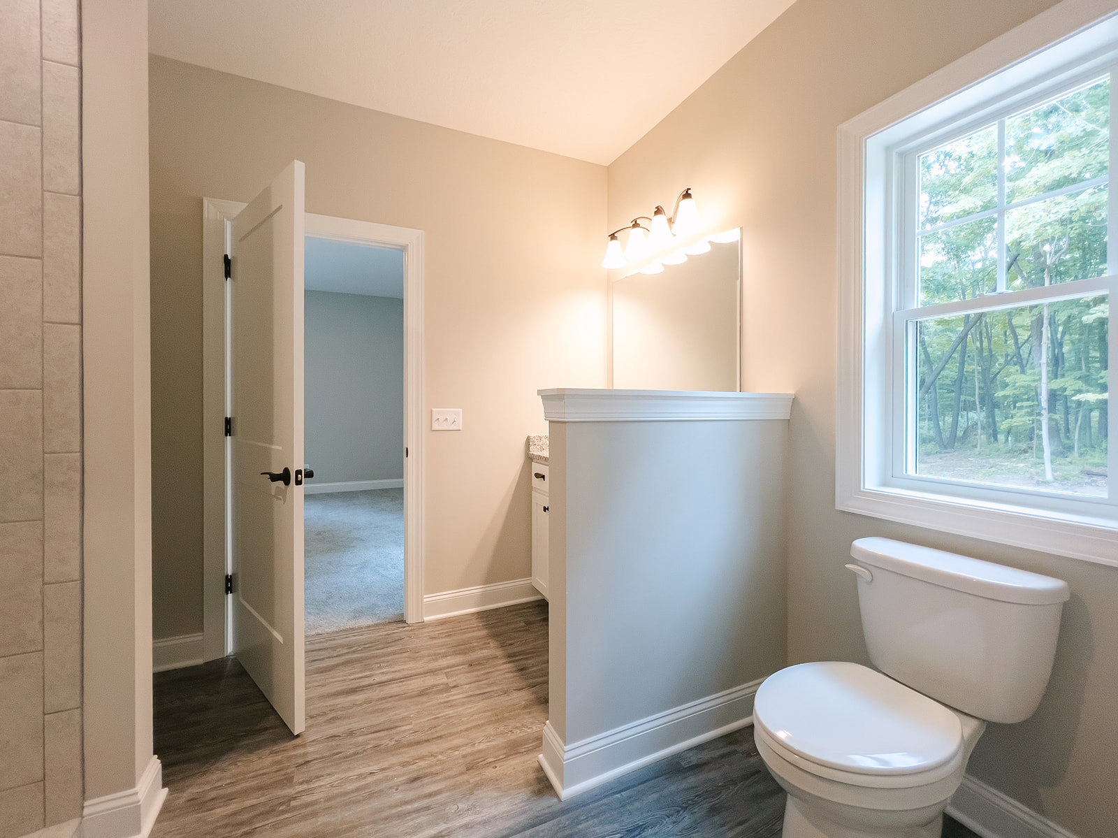 White toilet with closed lid beside a wall-mounted mirror, white tile walls, window showing trees outside, multi-bulb light fixture above mirror.