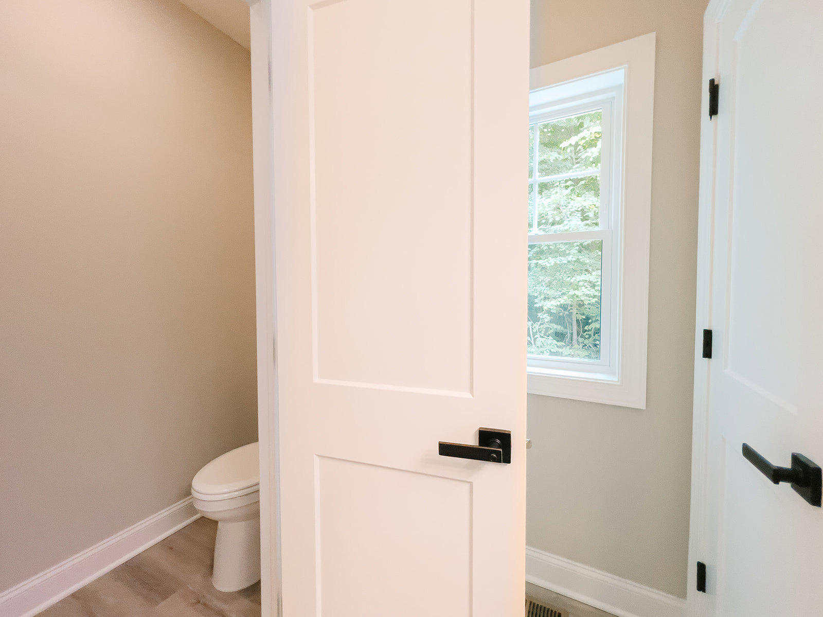 White paneled door with matte black handle, adjacent to a white-framed window and closed toilet, set against light plaster walls and neutral flooring in a bathroom.