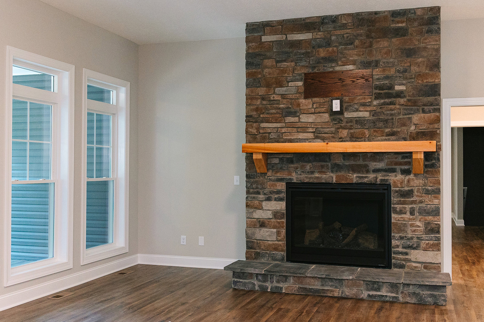 Brick fireplace with stacked logs, wood mantel shelf, exposed wood beam, glass-paned window, hardwood floor, and fire screen in a cozy den.