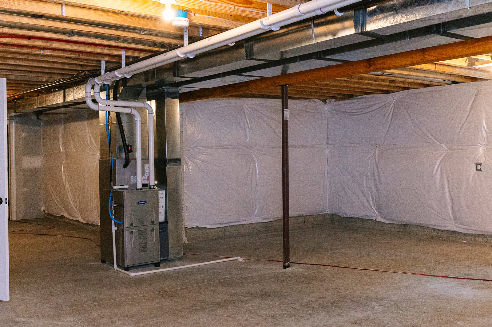 Basement utility room with exposed steel beams, grey machinery, blue hoses, and white sheet covering part of the floor.