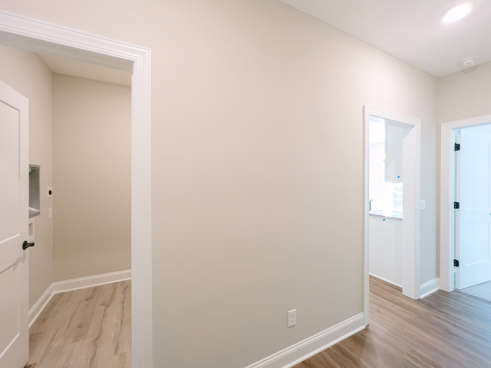 Hallway with white plaster walls, wood laminate flooring, white baseboards, white door with black handle, doorway, and window with blue objects visible outside