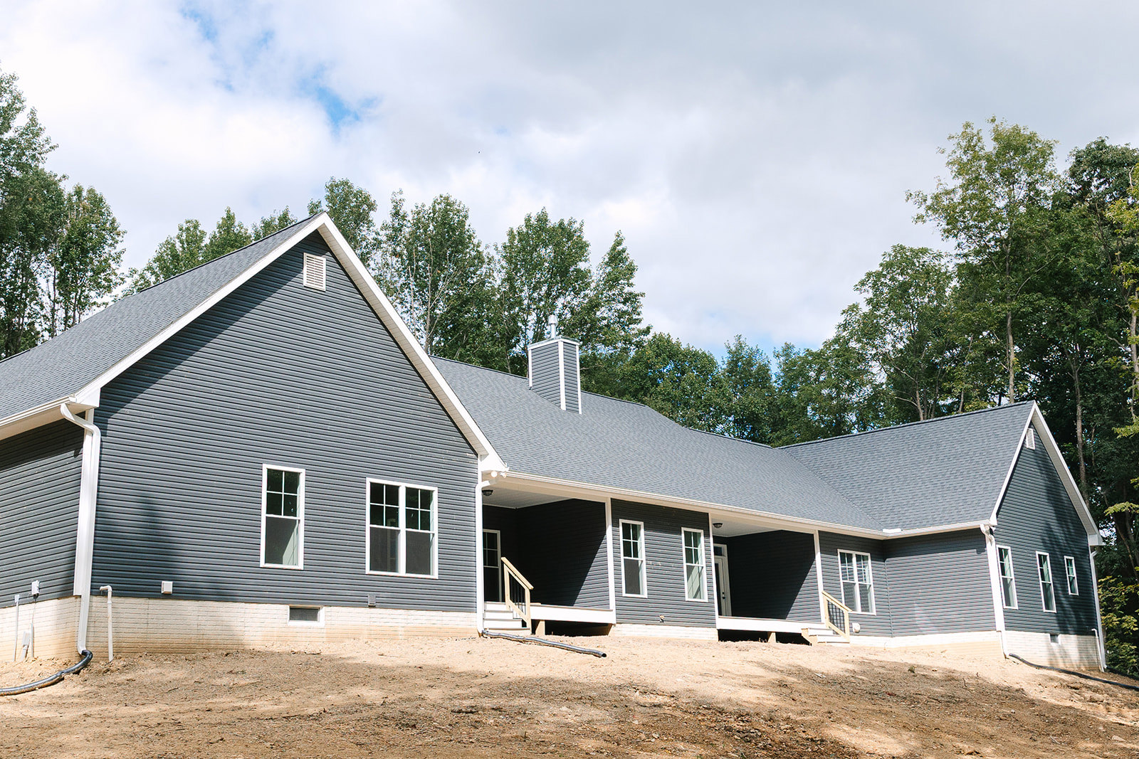 Two-story house surrounded by mature trees, large covered porch with white railings, white-framed windows, brick chimney, gray shingle roof, lush greenery in the background.