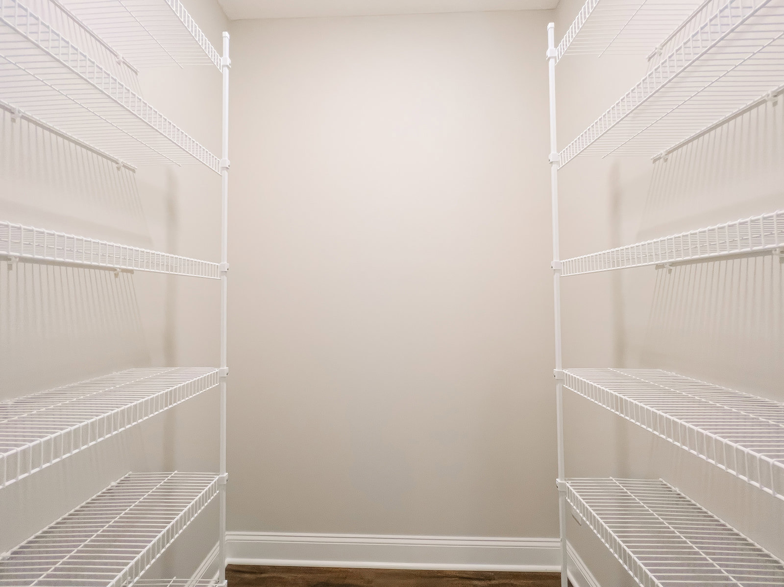 White metal wire shelving mounted on a white plaster wall, white ceiling with crown molding, brown hardwood floor with white baseboard.
