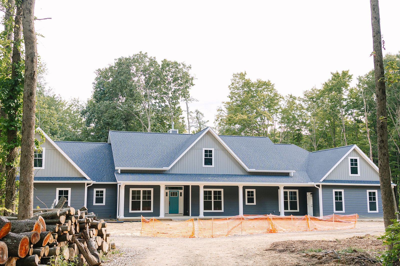 Partially built house with blue door, white trim, white pillars, and white-framed windows, surrounded by trees; pile of cut logs and close-up wooden fence in foreground