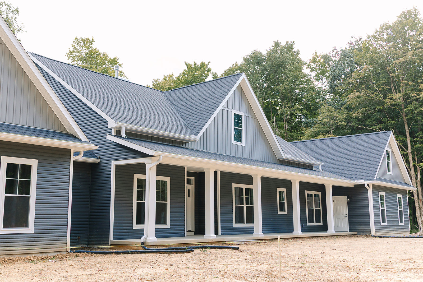 Spacious front porch with white framed windows, white door featuring black knobs, black pipe along ground, light siding, and mature trees in background