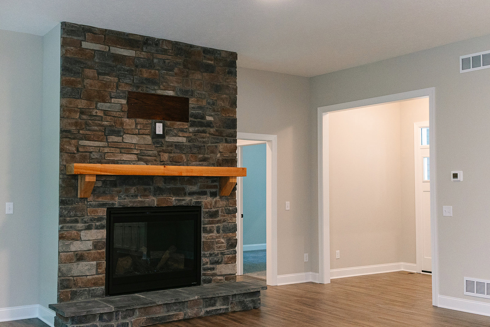 Stone hearth fireplace with glass door, wood mantel shelf above, white wall featuring light fixture and vent, hardwood floor.