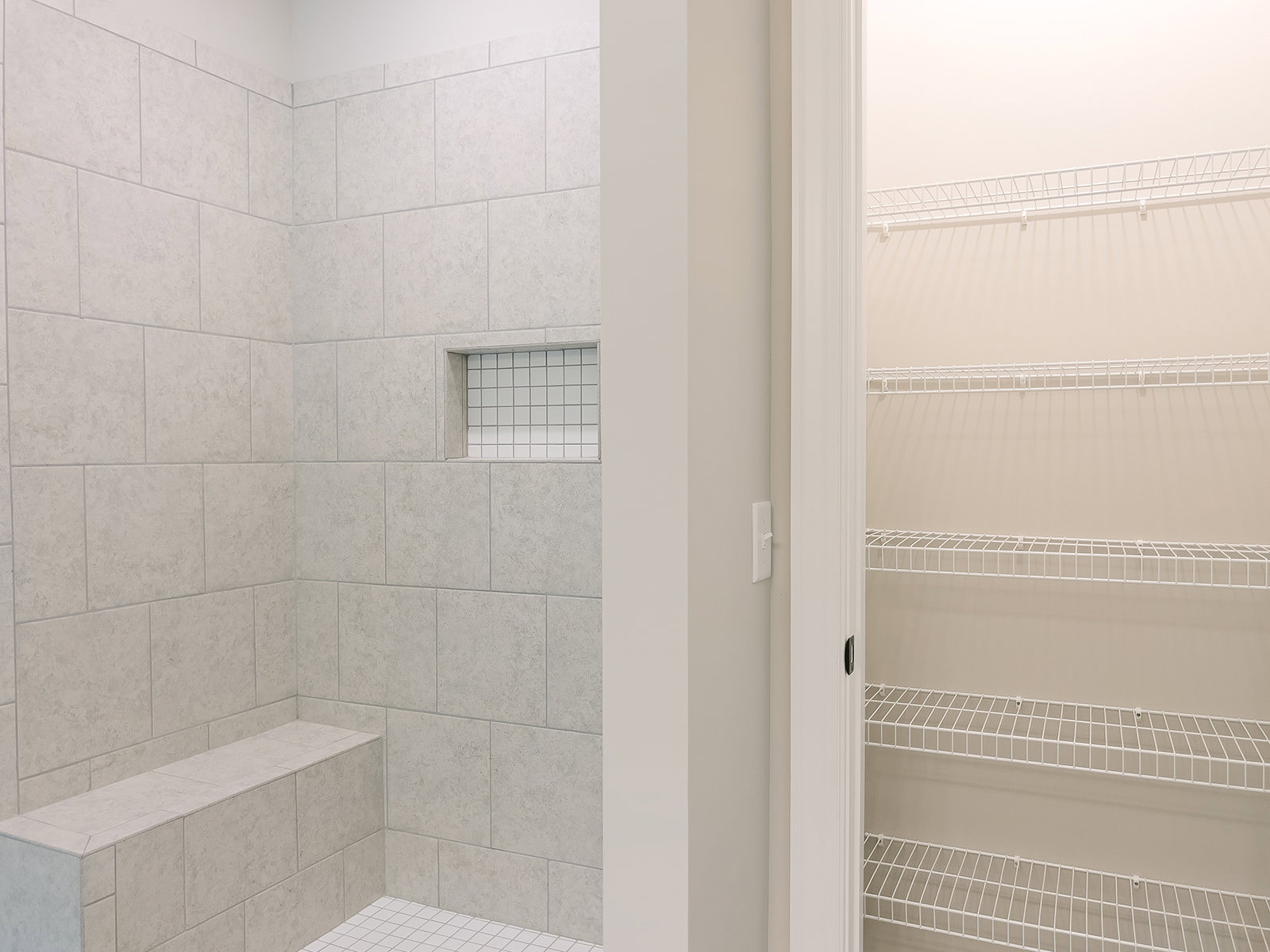 Walk-in shower with white tile walls, grey tile border, built-in stone bench, and recessed tile shelf; adjacent closet features white wire shelving