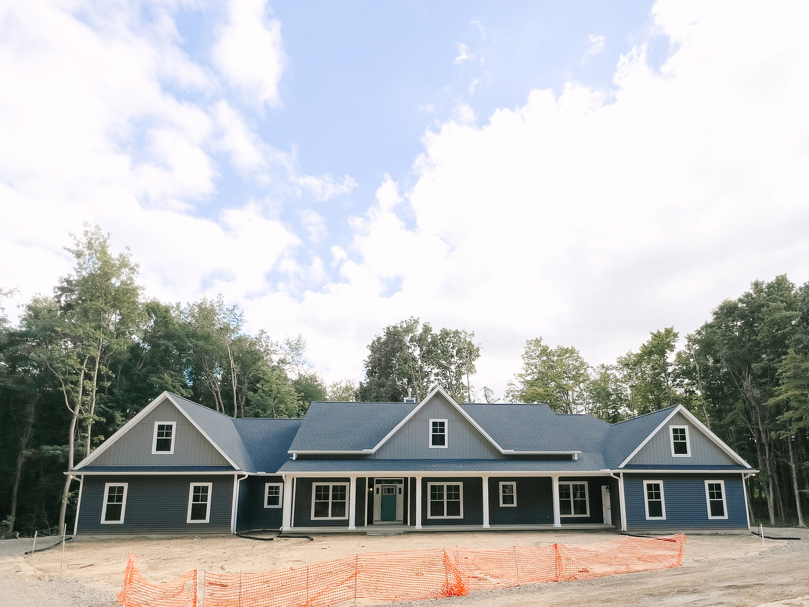 Framed house under construction with blue roof, white window frame, green door, surrounded by trees under partly cloudy sky