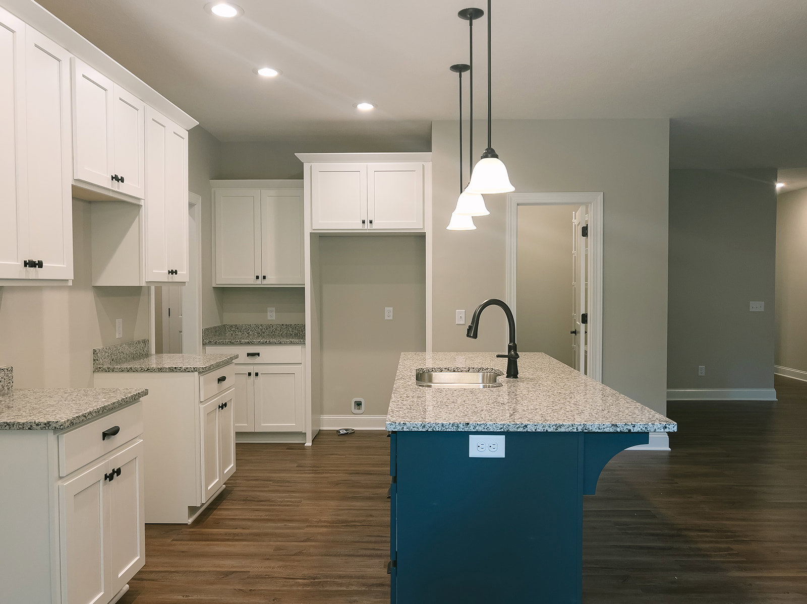 Granite kitchen island with undermount sink and black faucet, surrounded by white cabinets with black handles, light-colored flooring, and neutral walls