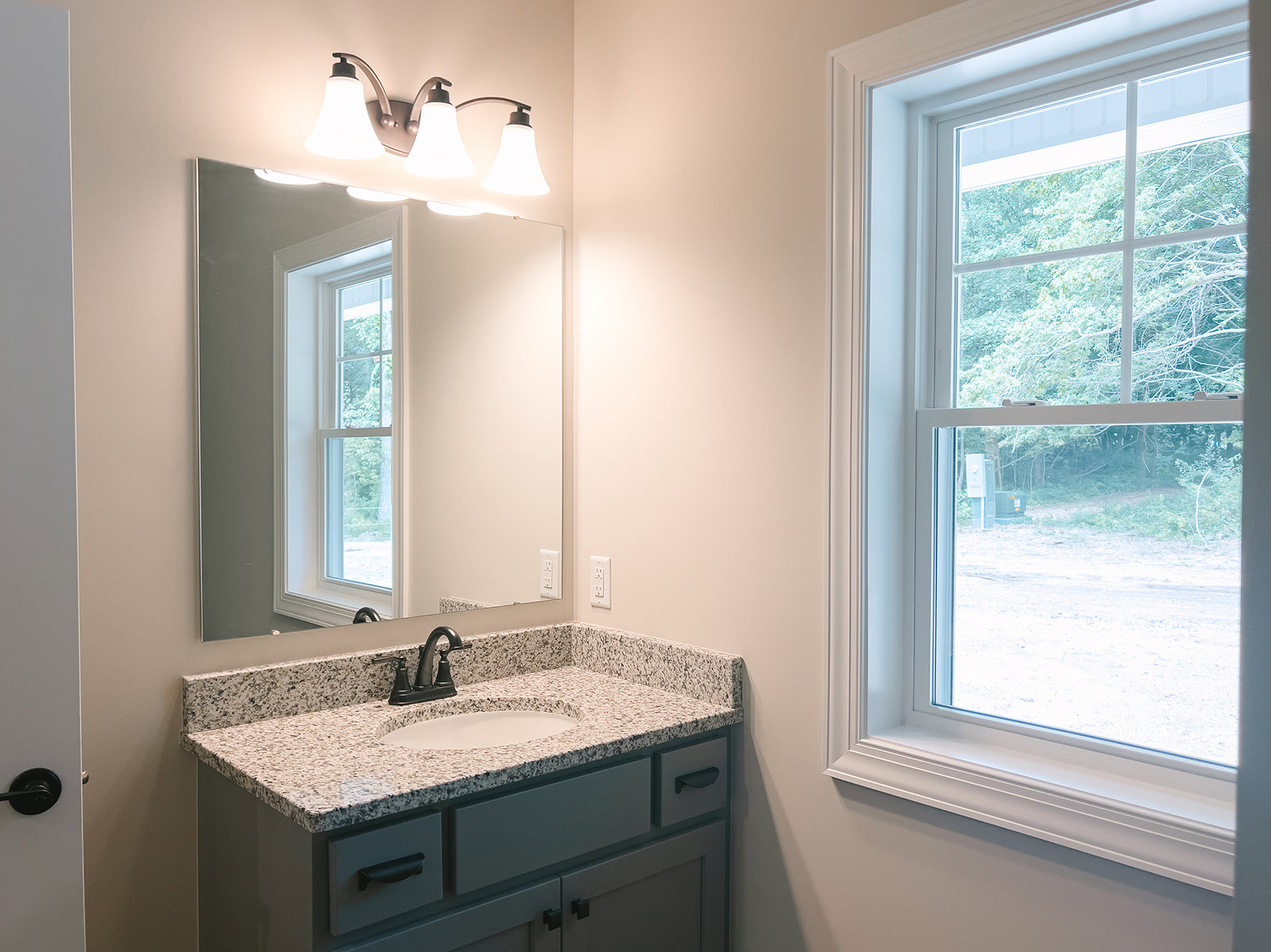 Bathroom with white tile walls, rectangular mirror above a vanity, black faucet on a white sink, three-light fixture mounted above the mirror, window with view of green trees
