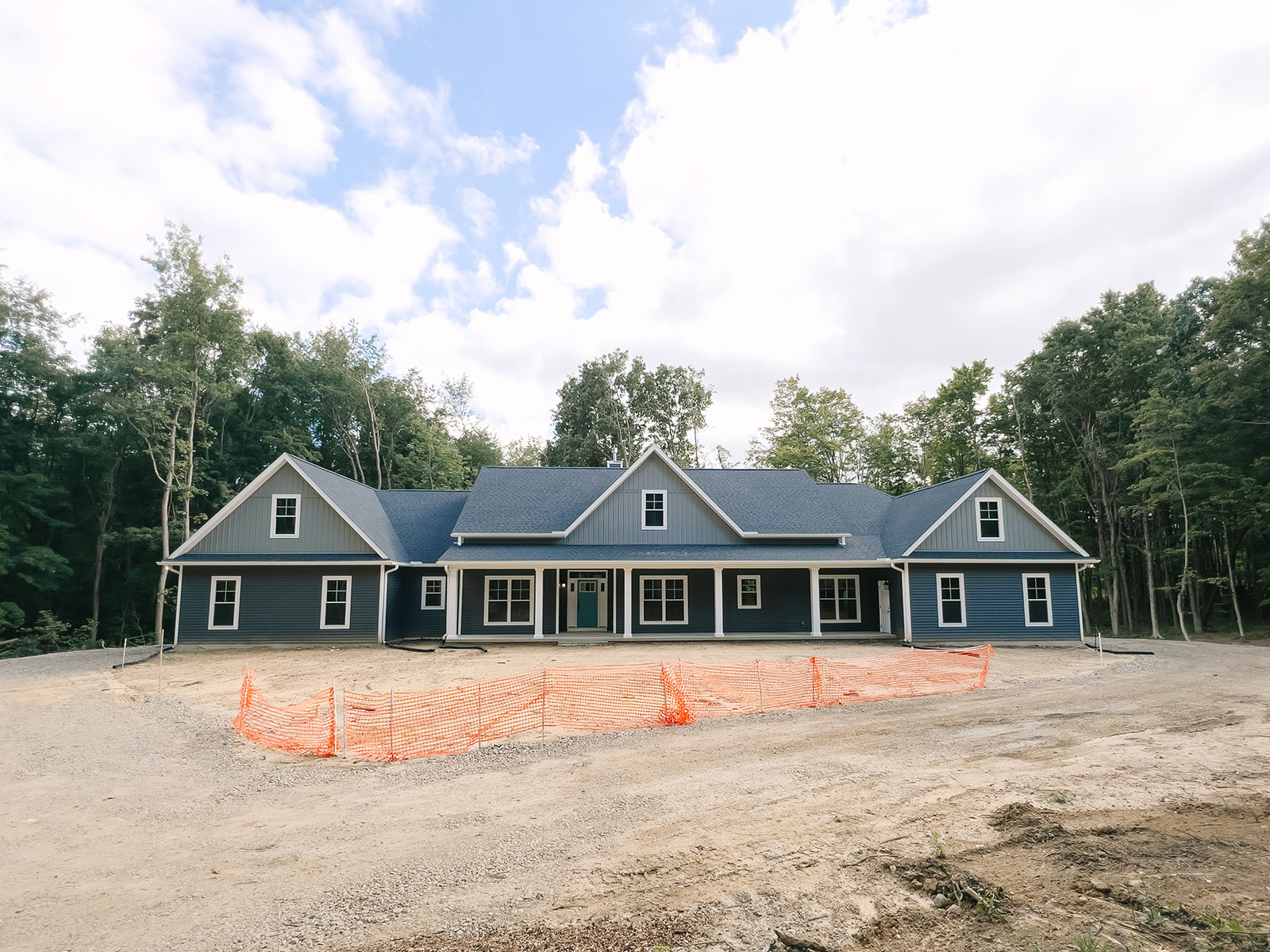 Two-story house under construction with white trim, blue front door featuring a window, orange construction mesh, white-framed windows, surrounded by trees under a partly cloudy