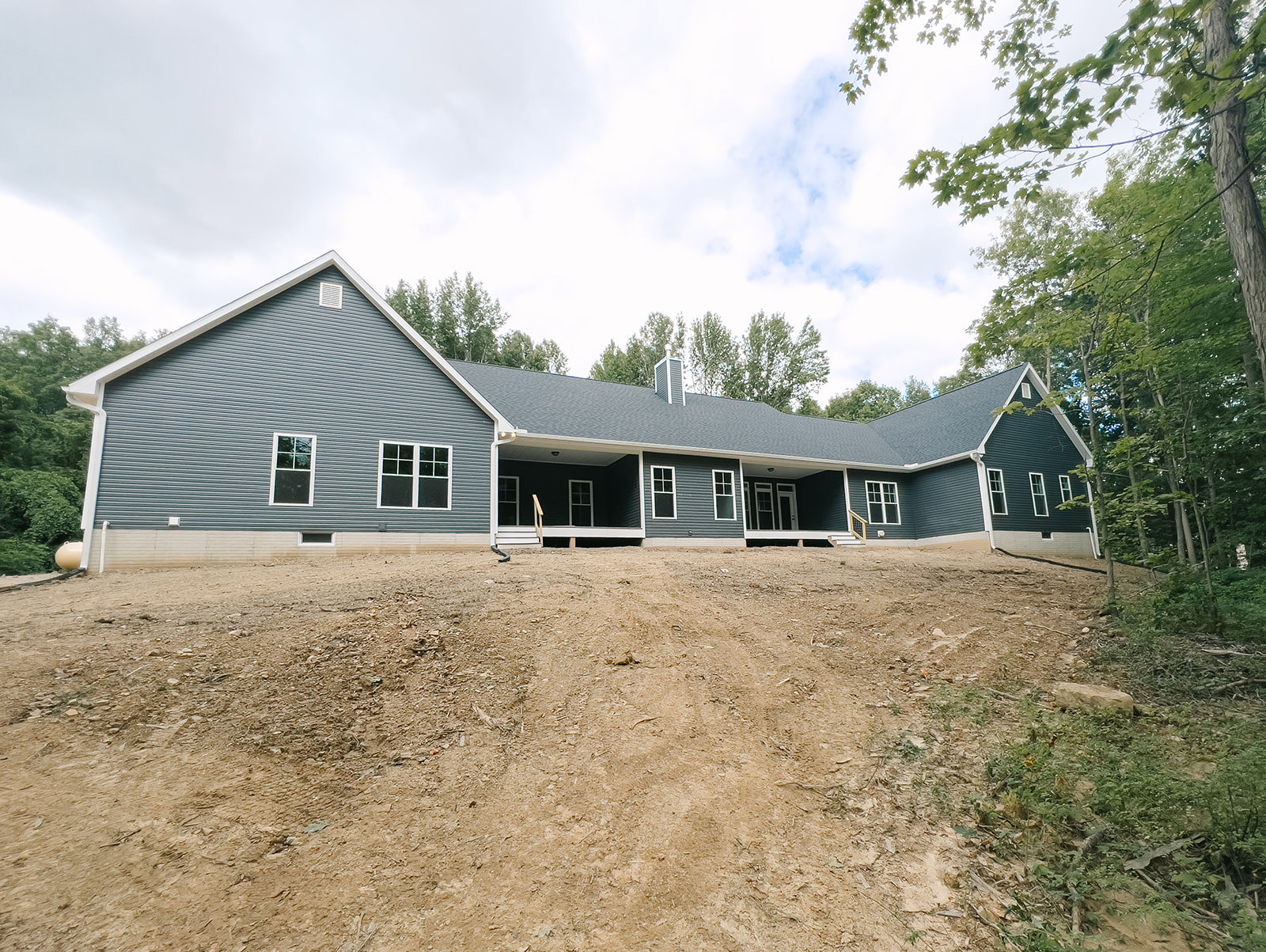 Two-story house with white-framed windows, covered porch, and exterior staircase, set behind a dirt hill under a cloudy sky.