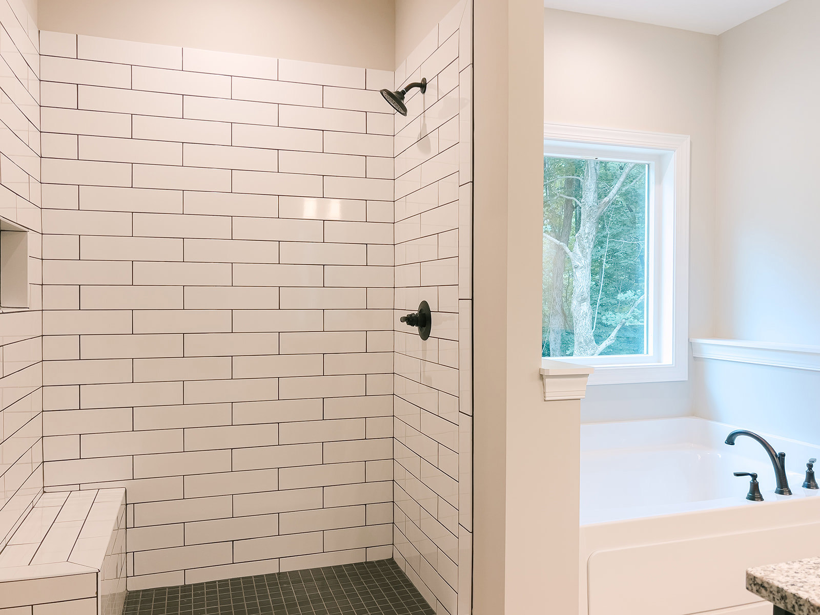Bathroom with freestanding tub and glass-enclosed shower, white tile walls, black tile floor, metal railing, tree visible through window, modern shower head, black fixture on white