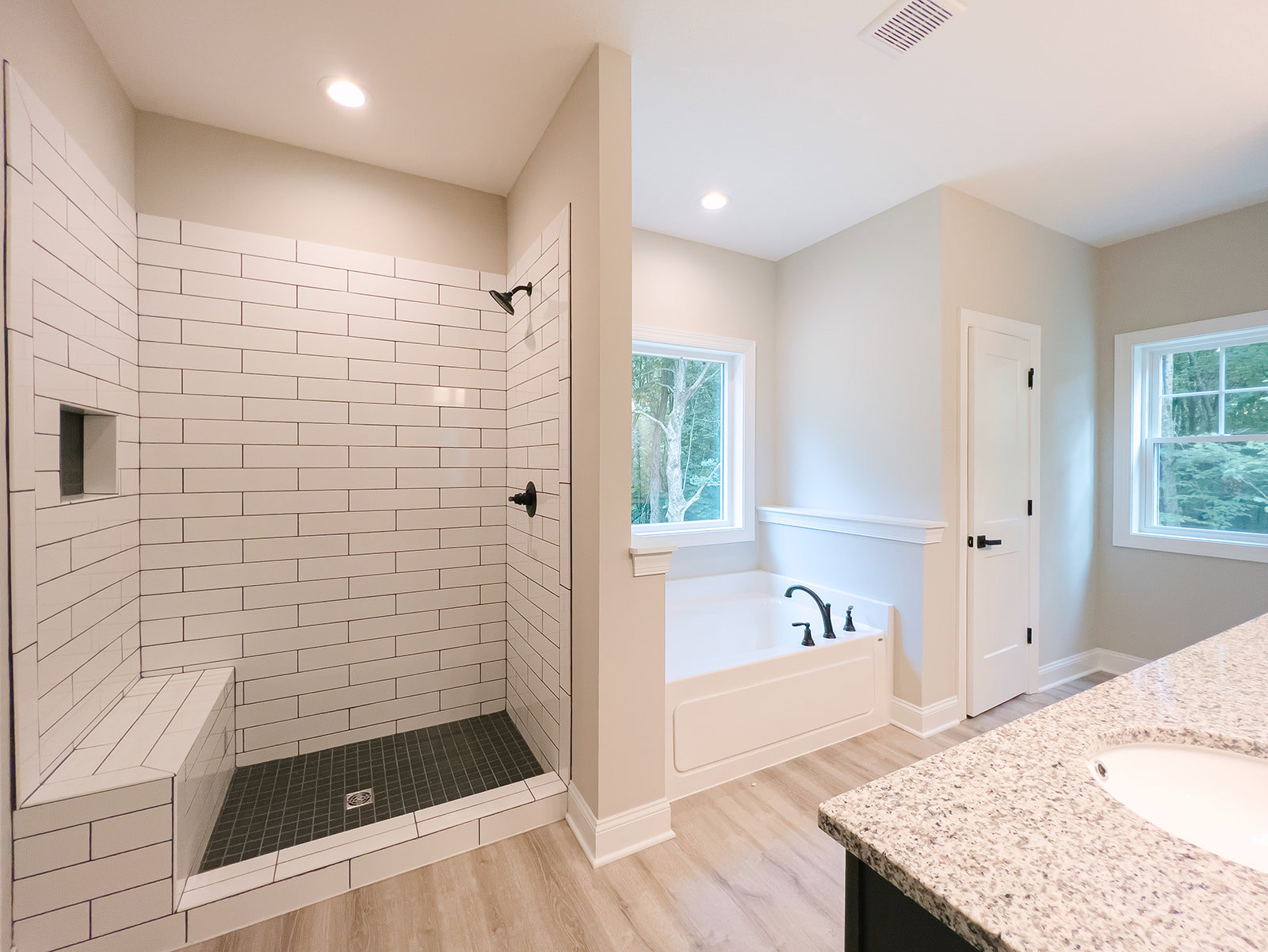 Bathroom with freestanding bathtub, rectangular sink on white vanity, large window with white frame showing leafy tree, white door with black handle, ceiling light, light gray tile