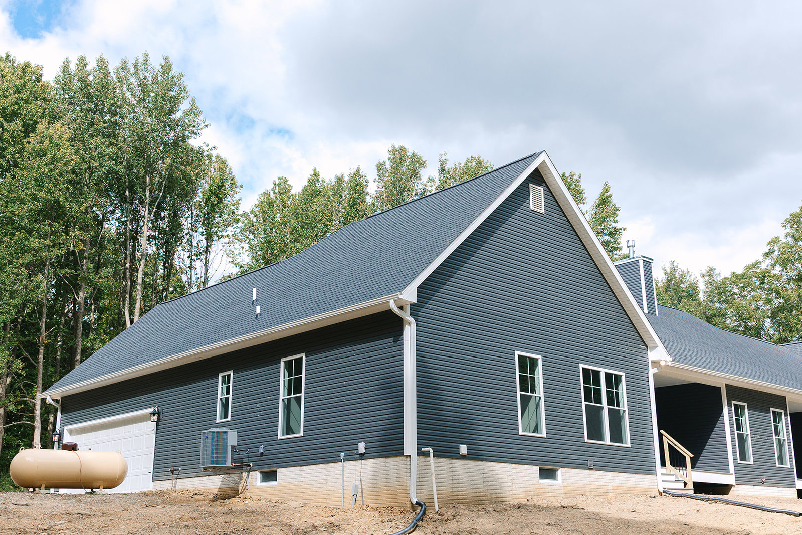 Two-story cottage with white-framed windows, gray roof, drainpipe, air conditioner unit, and water tank, surrounded by trees under a cloudy sky