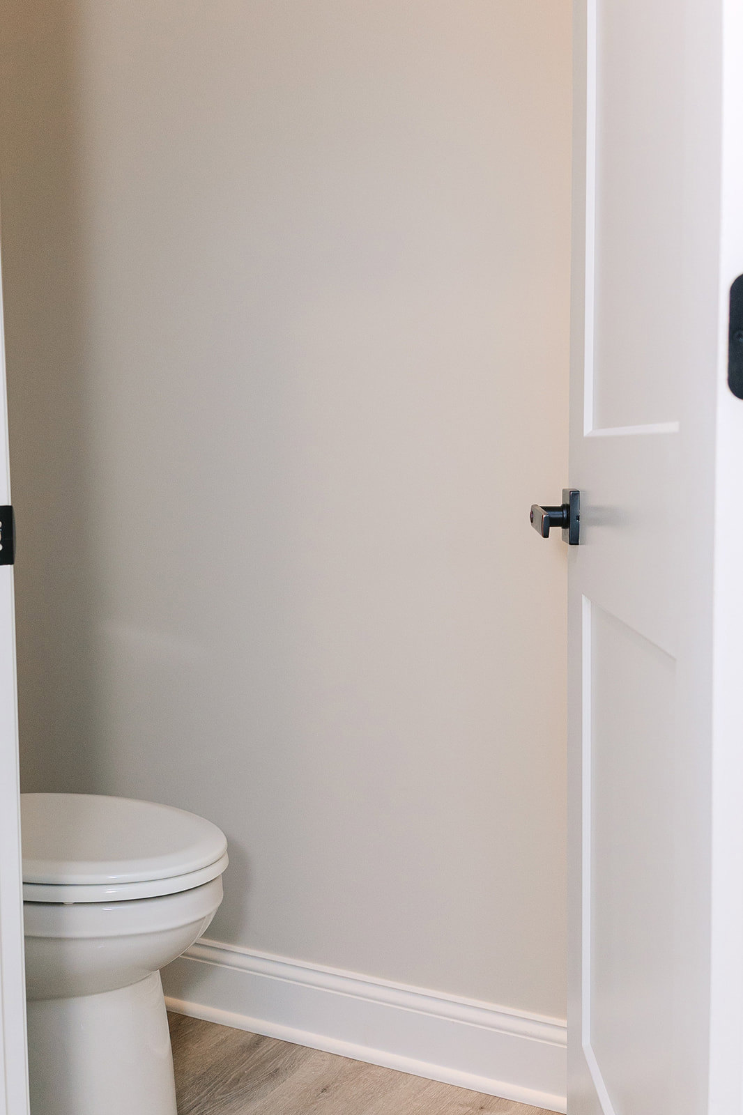 White door with matte black handle, white toilet with closed lid, wood flooring, and white walls in a modern bathroom.